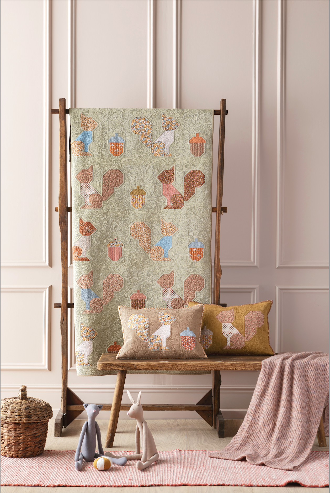 A quilted textile with a whimsical autumn pattern of squirrels, acorns, and leaves in muted earthy tones, displayed on a wooden stand in a styled nursery setting with matching pillows, a bench, and toys.
Photo by Tildafabrics.com [additional_image_link]