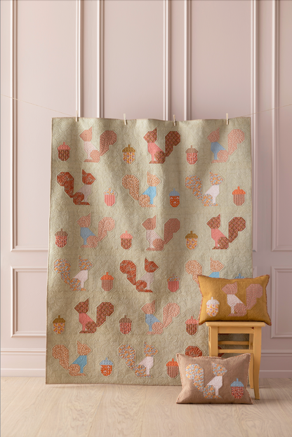 A beige fabric curtain or panel with a repeating pattern of stylized squirrels, acorns, and pumpkins in muted brown, pink, and light blue, hanging in a room with paneled walls. Two matching decorative pillows are placed on a small wooden stool and floor beside it.
Photo by Tildafabrics.com [additional_image_link]