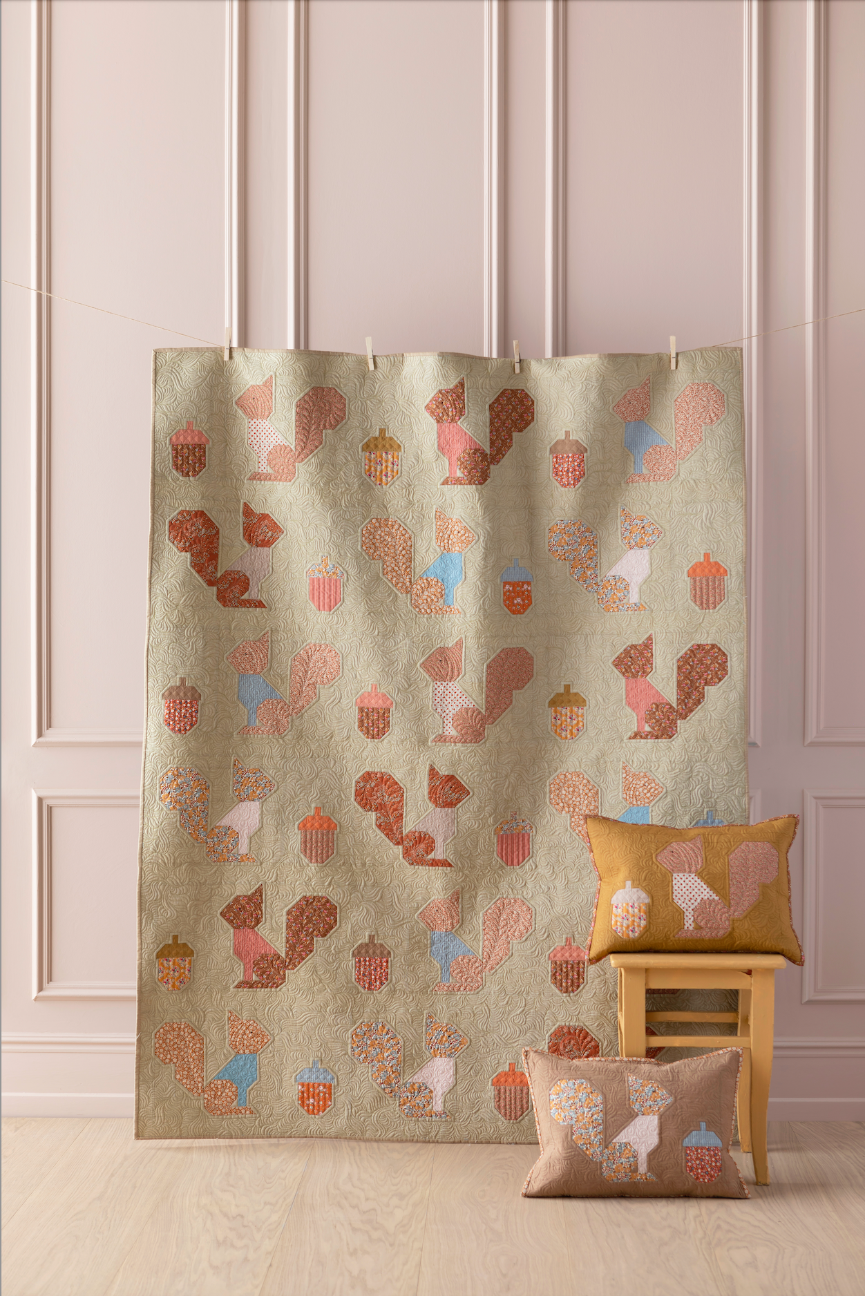 A beige fabric curtain or panel with a repeating pattern of stylized squirrels, acorns, and pumpkins in muted brown, pink, and light blue, hanging in a room with paneled walls. Two matching decorative pillows are placed on a small wooden stool and floor beside it.
Photo by Tildafabrics.com [additional_image_link]