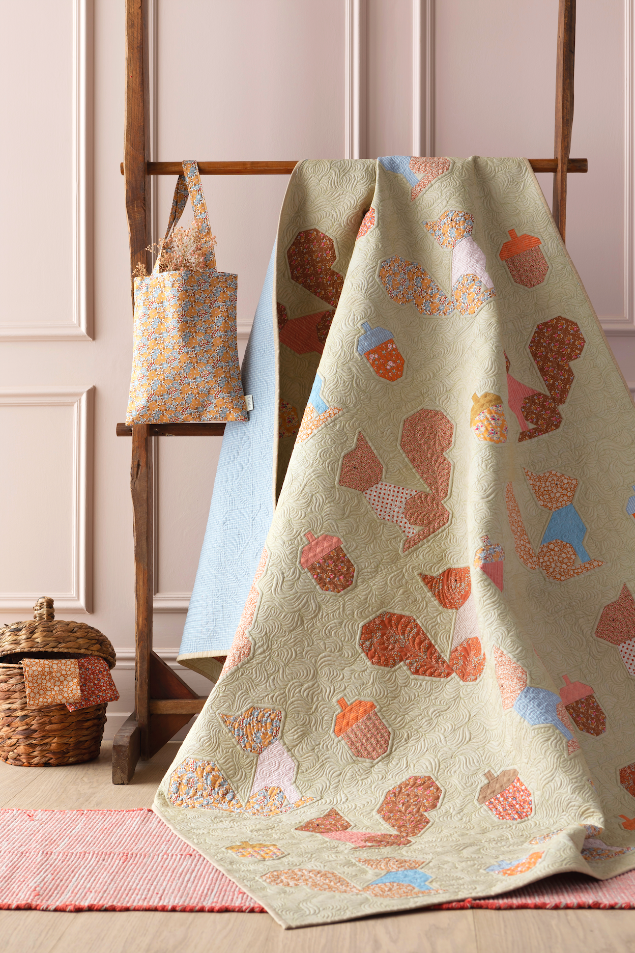 A quilted blanket with a patchwork acorn pattern in muted earthy tones, draped over a wooden ladder rack in a styled room setting.
Photo by Tildafabrics.com [additional_image_link]