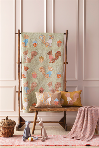 A quilted textile with a whimsical autumn pattern of squirrels, acorns, and leaves in muted earthy tones, displayed on a wooden stand in a styled nursery setting with matching pillows, a bench, and toys.
Photo by Tildafabrics.com [additional_image_link]