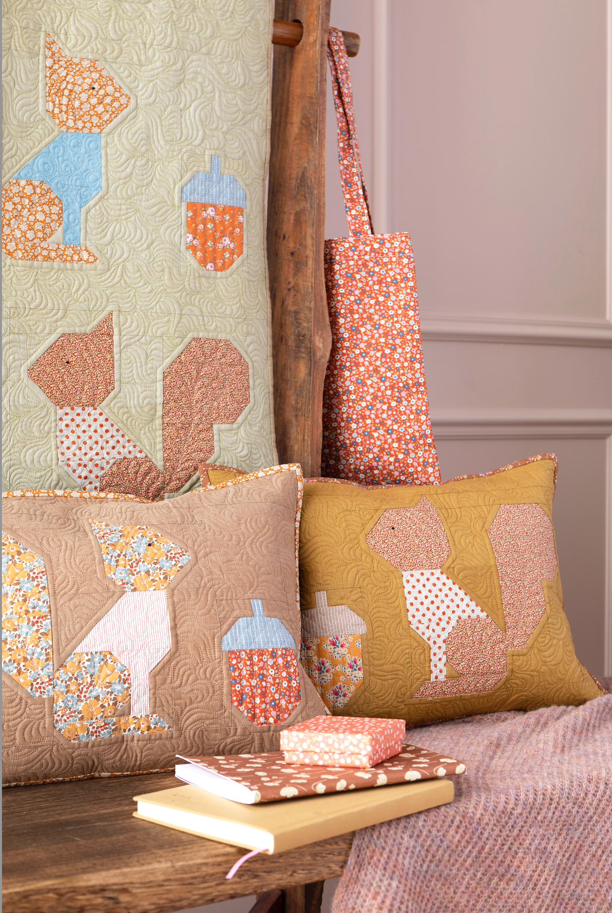 A styled home decor scene featuring two quilted throw pillows with fox and acorn motifs, a matching quilted fabric panel, a patterned tote bag hanging on a wooden chair, and a stack of books on a bench.
Photo by Tildafabrics.com [additional_image_link]
