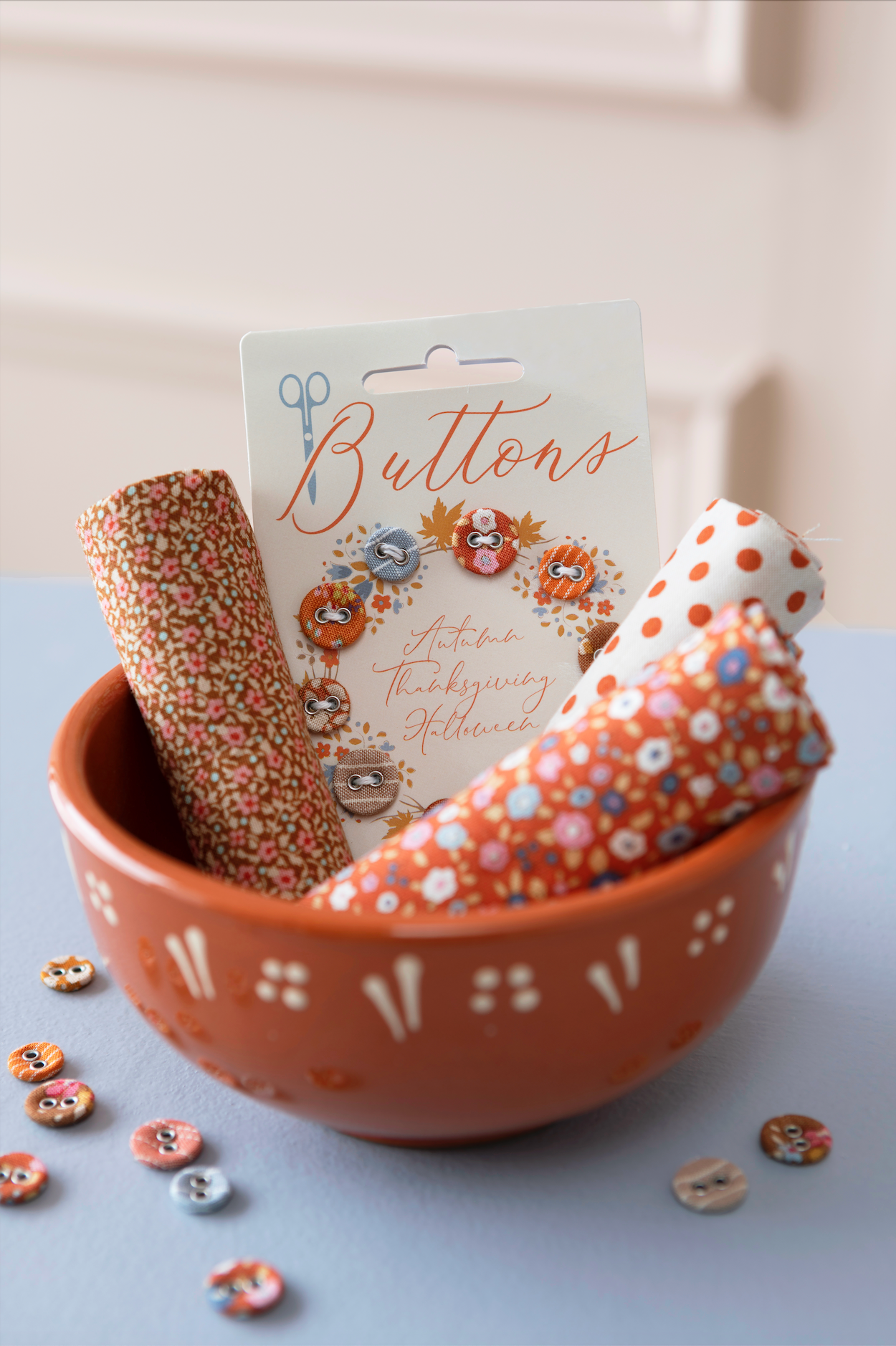 A terracotta-colored ceramic bowl filled with decorative buttons and button rolls, with a hanging card labeled 'Buttons' and autumn-themed illustrations. The scene is styled on a pale blue surface against a soft cream background.
Photo by Tildafabrics.com [additional_image_link]
