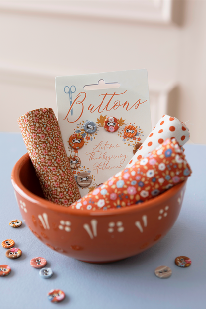 A terracotta-colored ceramic bowl filled with decorative buttons and button rolls, with a hanging card labeled 'Buttons' and autumn-themed illustrations. The scene is styled on a pale blue surface against a soft cream background.
Photo by Tildafabrics.com [additional_image_link]