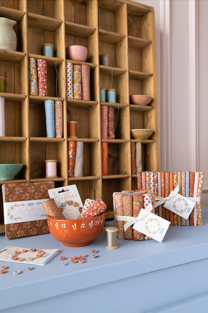 A styled interior scene showing a light blue mantel or shelf with folded fabrics, patterned paper goods, a terracotta bowl, and cards, set in front of a large wooden cubby shelving unit filled with matching home decor items.
Photo by Tildafabrics.com [additional_image_link]