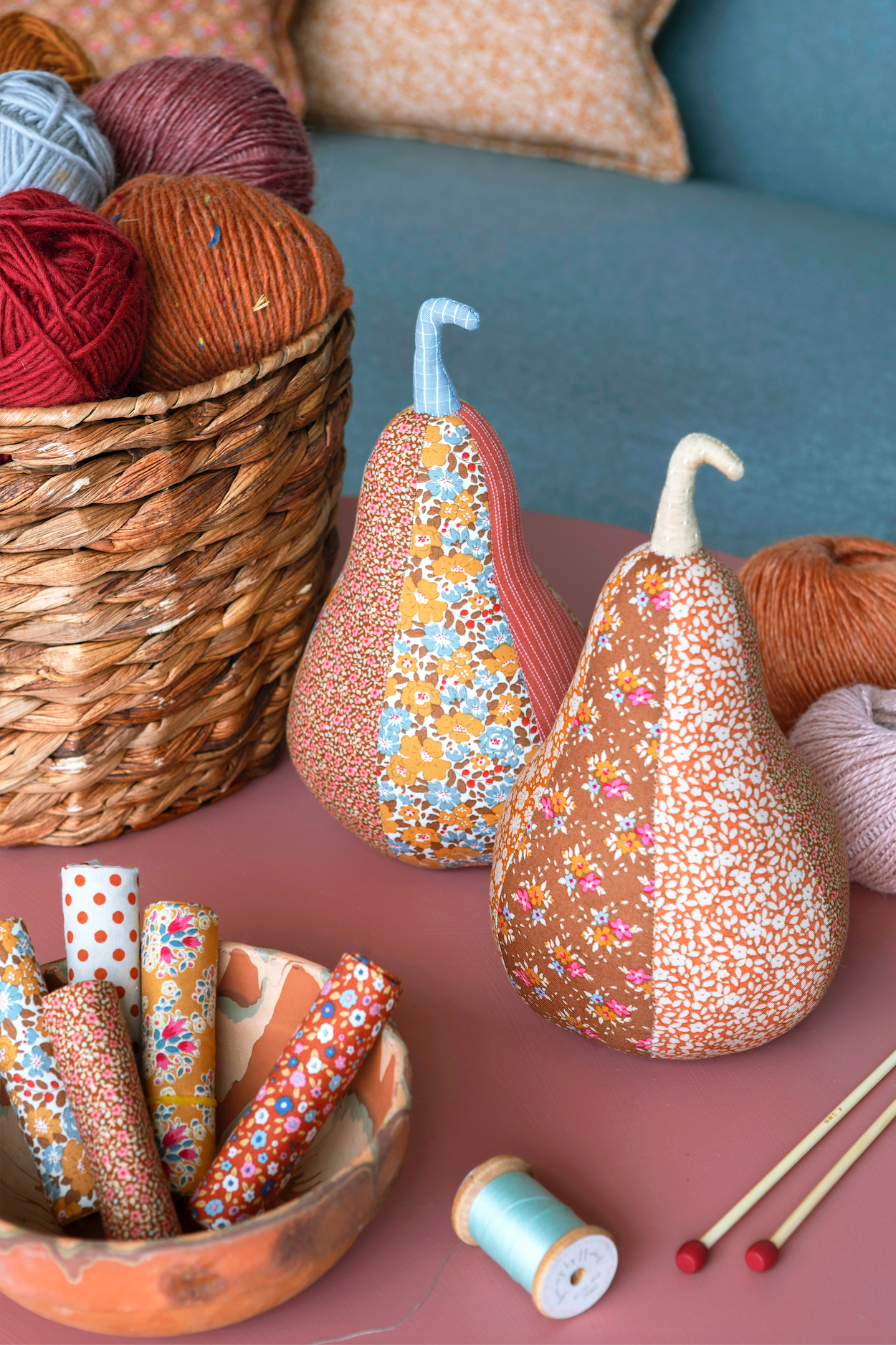 Two decorative pear-shaped fabric ornaments with floral patterns and stitched stems, displayed on a pink table alongside yarn, thread rolls, knitting needles, and a wicker basket in a cozy craft setting.
Photo by Tildafabrics.com [additional_image_link]