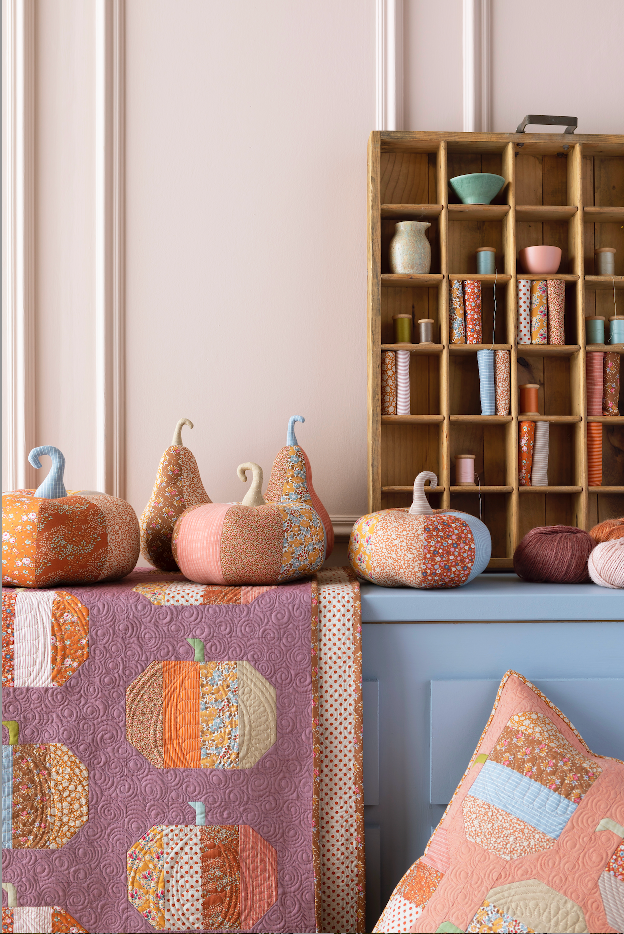 A styled interior scene with multiple handmade patchwork pumpkins and gourds arranged on a table and shelf, alongside a quilted blanket and matching pillows in warm autumn tones. A wooden bookcase stands in the background against a pale pink wall.
Photo by Tildafabrics.com [additional_image_link]