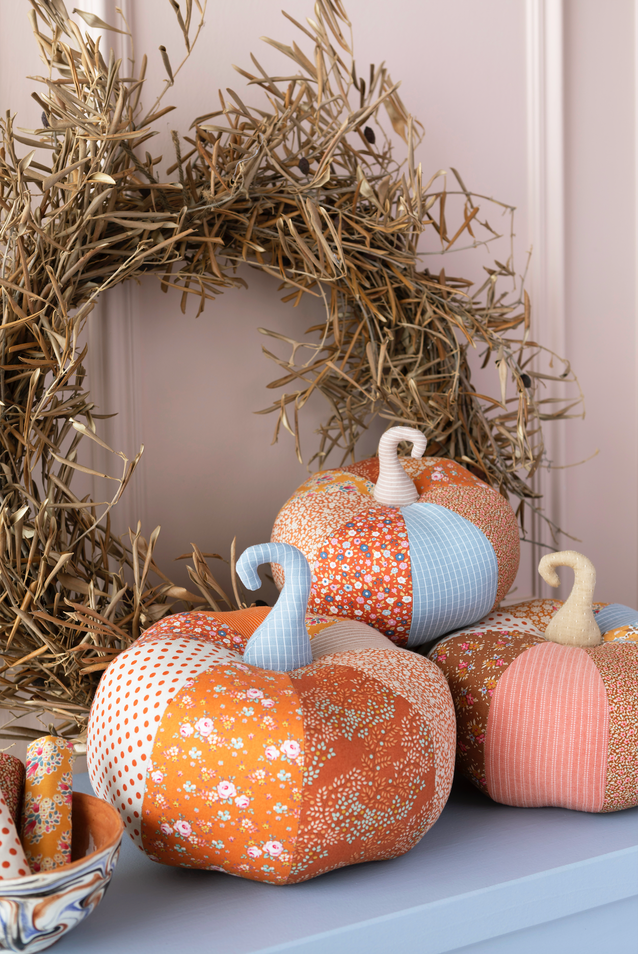 A group of handmade patchwork fabric pumpkins with faux stems arranged on a light blue shelf in front of a dried straw wreath.
Photo by Tildafabrics.com [additional_image_link]