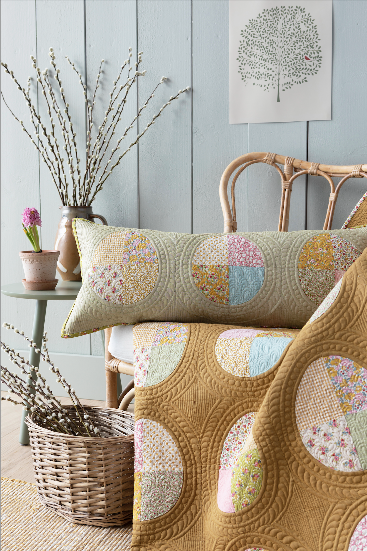 A quilted blanket and matching lumbar pillow featuring circular patchwork fabric panels in soft pastel colors, styled on a rattan chair in a rustic room setting with a blue paneled wall, side table, and basket.
Photo by Tildafabrics.com [additional_image_link]