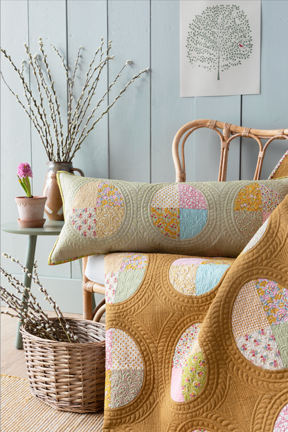 A quilted blanket and matching lumbar pillow featuring circular patchwork fabric panels in soft pastel colors, styled on a rattan chair in a rustic room setting with a blue paneled wall, side table, and basket.
Photo by Tildafabrics.com [additional_image_link]