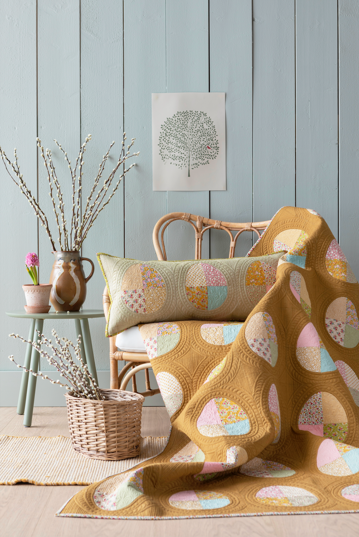 A quilted blanket and matching pillow with a pastel egg pattern draped over a rattan chair in a styled room with light blue paneled walls, a side table, and decorative branches.
Photo by Tildafabrics.com [additional_image_link]
