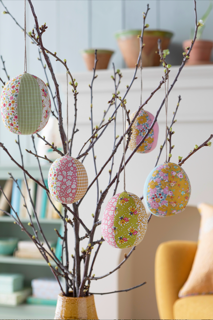 Decorative hanging Easter eggs with floral and checkered fabric patterns displayed on a branch arrangement in a vase, set in a softly blurred indoor room.
Photo by Tildafabrics.com [additional_image_link]