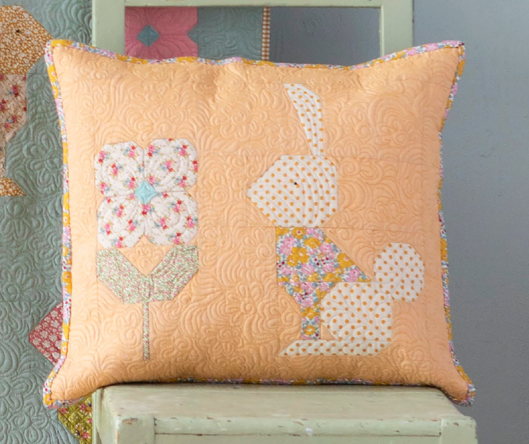 A square quilted throw pillow in peach with applique patches of a rabbit and a flower, resting on a distressed white chair.
Photo by Tildafabrics.com [additional_image_link]