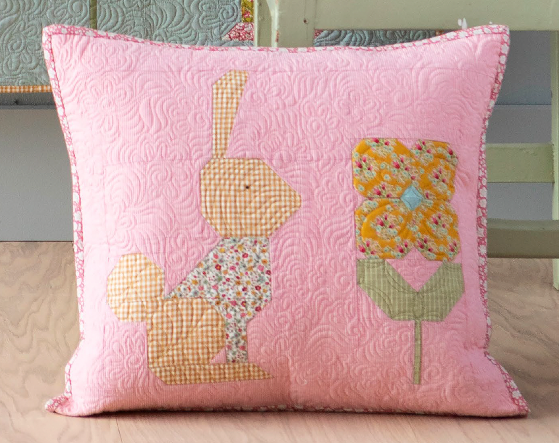 A square pink quilted throw pillow featuring an applique design of a bunny and a flower, shown resting on a light wood floor.
Photo by Tildafabrics.com [additional_image_link]