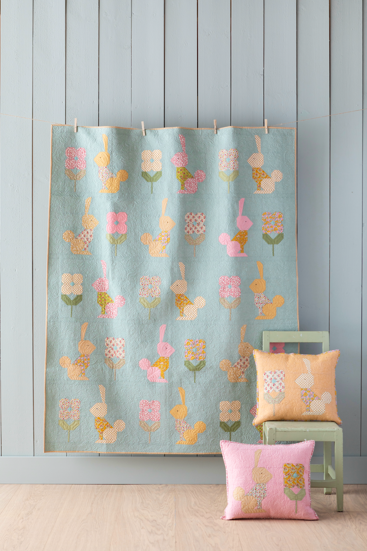 A quilted fabric panel with a repeating pattern of bunnies, squirrels, and flowers in pastel colors, hanging on a light blue-gray paneled wall. A small green chair and two matching decorative pillows are placed below it on a light wood floor.
Photo by Tildafabrics.com [image_link]