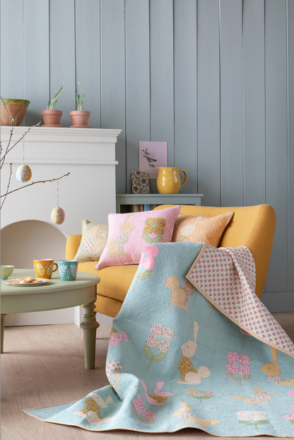 A light blue quilted throw blanket with a whimsical bunny and floral pattern is draped over a mustard yellow armchair in a styled living room. The scene includes coordinating pink and yellow pillows, a light green coffee table with colorful mugs, and a white fireplace mantel against a blue paneled wall.
Photo by Tildafabrics.com [additional_image_link]