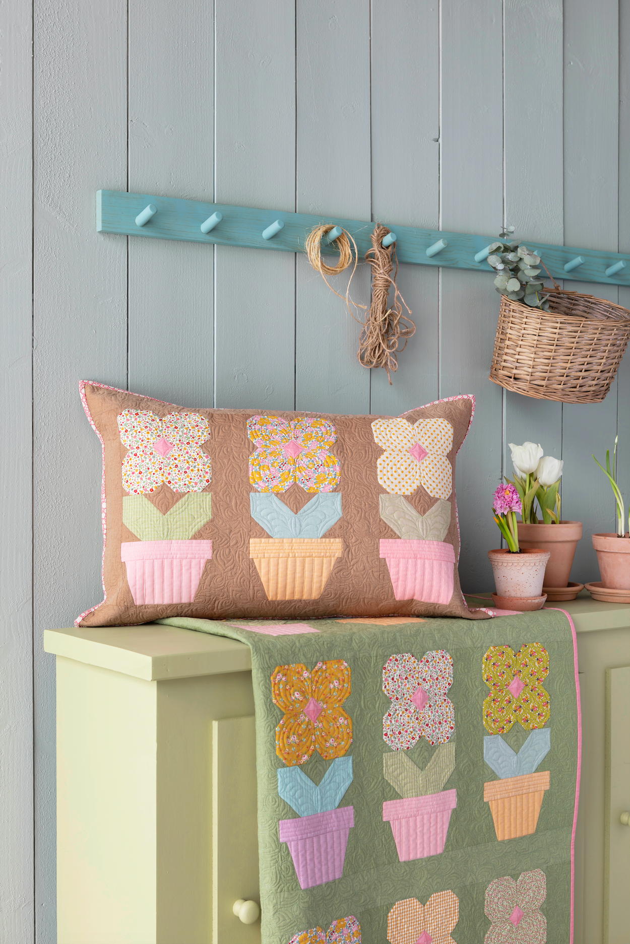 A decorative rectangular throw pillow with a floral pot pattern in muted pastel colors, resting on a pale green cabinet next to a matching quilted textile. The background features a light blue-gray paneled wall with a rustic teal peg rail, hanging twine, and a small wicker basket with potted flowers.
Photo by Tildafabrics.com [image_link]