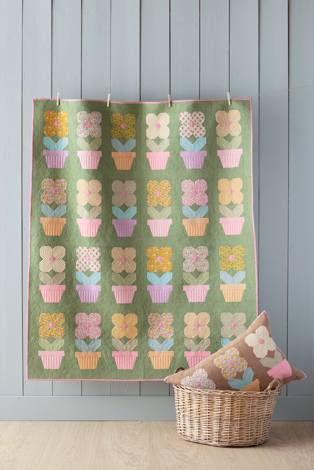 A quilted wall hanging or tapestry with a grid of pastel flower-botique motifs in pink, yellow, blue, and cream on an olive green background, displayed against a light gray paneled wall above a wicker basket with pillows.
Photo by Tildafabrics.com [additional_image_link]