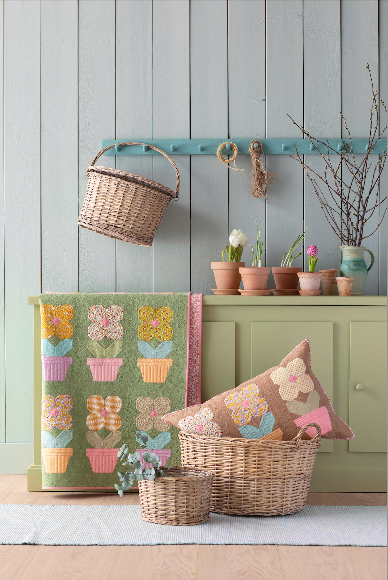 A styled home decor scene featuring a green quilted patchwork floral blanket with flower-pot motifs, a matching floral throw pillow on a wicker basket, a small wicker basket, and a sage green cabinet against a gray wood-paneled wall.
Photo by Tildafabrics.com [additional_image_link]