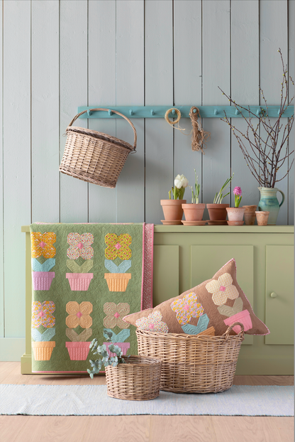 A styled home decor scene featuring a green quilted patchwork floral blanket with flower-pot motifs, a matching floral throw pillow on a wicker basket, a small wicker basket, and a sage green cabinet against a gray wood-paneled wall.
Photo by Tildafabrics.com [additional_image_link]