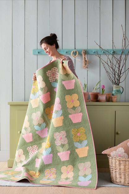 A woman holds up a large quilted blanket featuring a green background with colorful flowers in pots motifs, set against a rustic indoor background with a green cabinet and light gray wood-paneled wall.
Photo by Tildafabrics.com [additional_image_link]