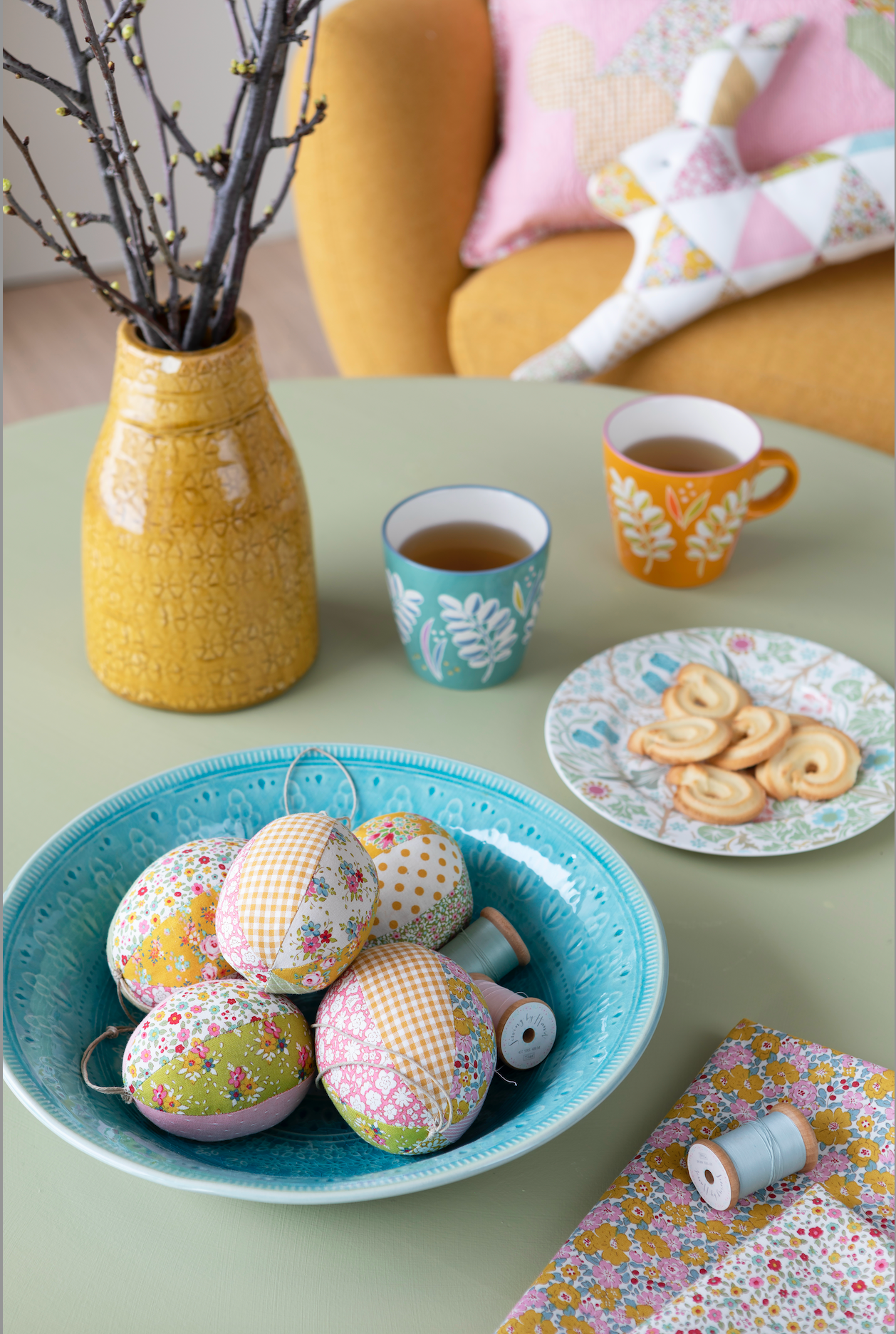 A styled tabletop scene with fabric-covered decorative eggs in a turquoise bowl, two patterned mugs, cookies on a plate, and a yellow vase with branches in the background. No person or animal is present, but this is not a clean isolated studio product shot; it is a real interior setting, so subject_type is Lifestyle.
Photo by Tildafabrics.com [additional_image_link]