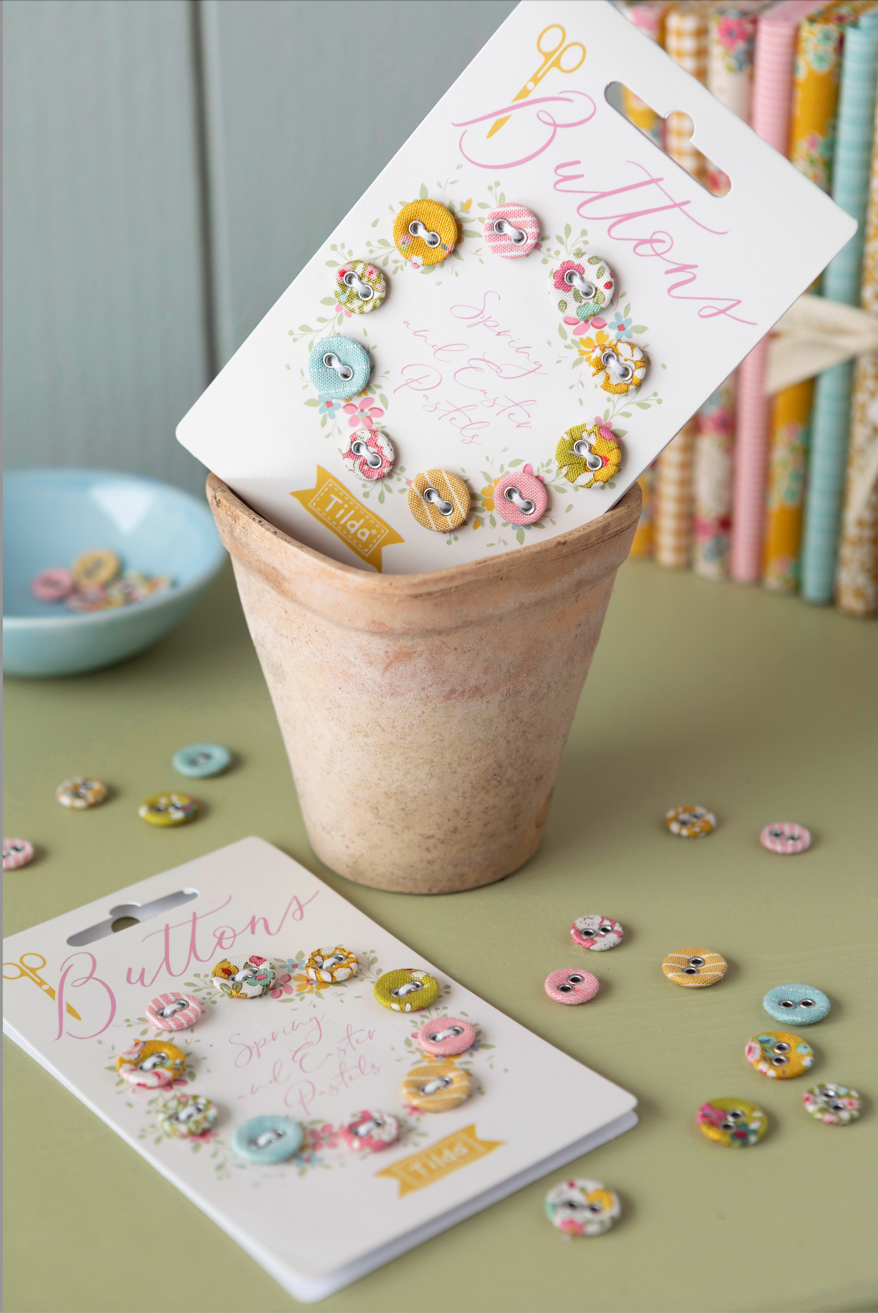 Two white display cards labeled 'Buttons' with assorted pastel decorative buttons, arranged in and around a small beige pot on a muted green surface, with scattered loose buttons and a bookshelf in the background.
Photo by Tildafabrics.com [additional_image_link]