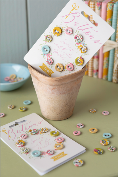 Two white display cards labeled 'Buttons' with assorted pastel decorative buttons, arranged in and around a small beige pot on a muted green surface, with scattered loose buttons and a bookshelf in the background.
Photo by Tildafabrics.com [additional_image_link]