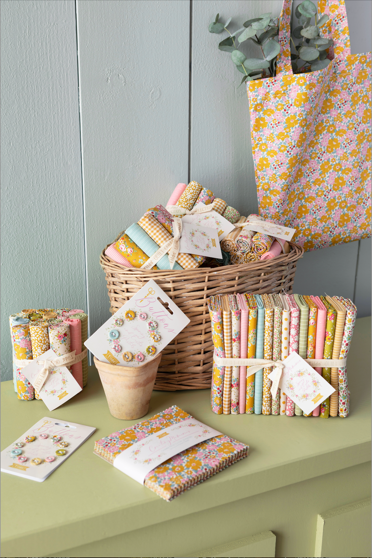 A styled collection of fabric quilts and a matching fabric tote bag in a soft yellow and pink floral print, arranged on a light green table against a gray paneled wall.
Photo by Tildafabrics.com [additional_image_link]