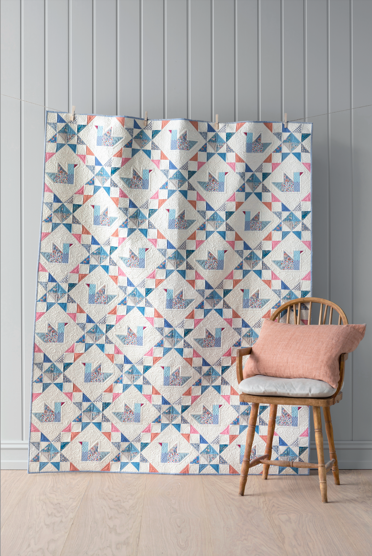 A large quilt featuring a repeating geometric patchwork pattern in blue, pink, and gray on a white background (ducks), displayed in a room setting next to a wooden chair with a pink cushion.
Photo by Tildafabrics.com [image_link]