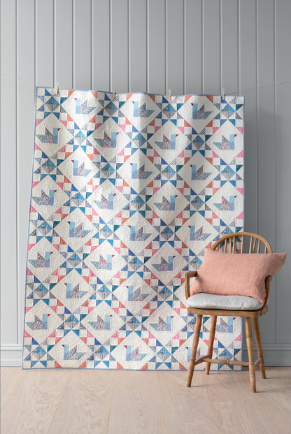 A large quilt featuring a repeating geometric patchwork pattern in blue, pink, and gray on a white background (ducks), displayed in a room setting next to a wooden chair with a pink cushion.
Photo by Tildafabrics.com [image_link]