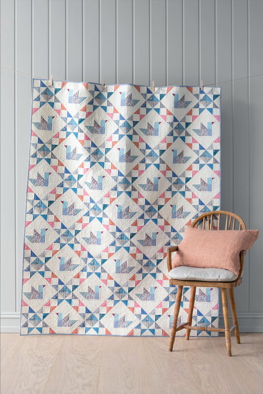 A large quilt featuring a repeating geometric patchwork pattern in blue, pink, and gray on a white background (ducks), displayed in a room setting next to a wooden chair with a pink cushion.
Photo by Tildafabrics.com [image_link]