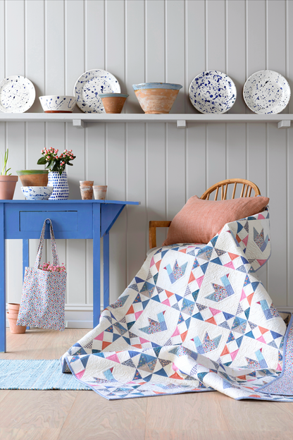 A quilted throw blanket with a geometric bird pattern in blue, pink, and gray on a white background, draped over a wooden chair in a styled room setting.
Photo by Tildafabrics.com [additional_image_link]