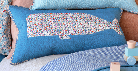 A blue quilted rectangular throw pillow featuring a whale patchwork filled with a multicolor floral pattern, styled on a bed with other blue pillows and blankets.
Photo by Tildafabrics.com [image_link]