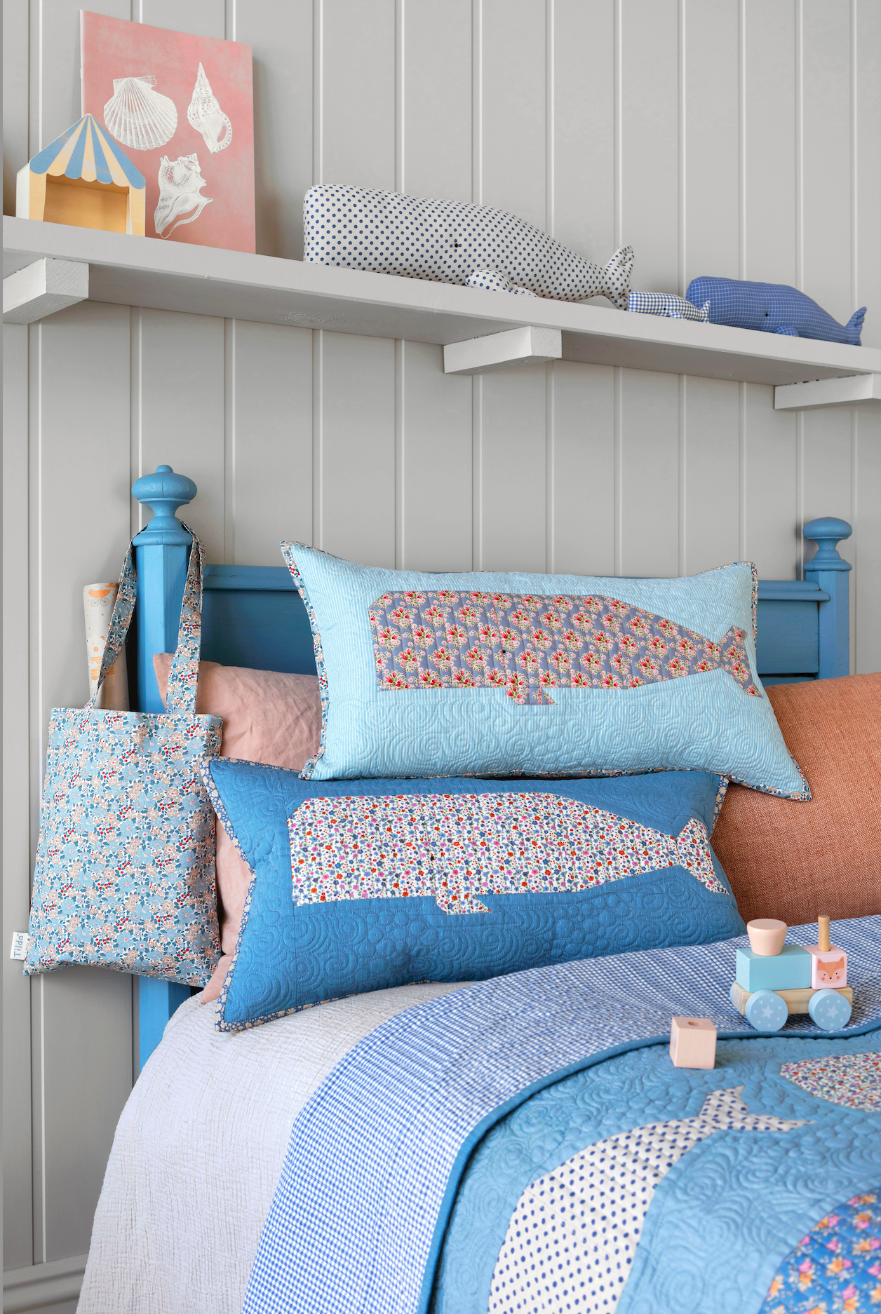A styled children's bedroom scene showing a bed with blue bedding, whale-patterned pillows, a blue wooden headboard, wall shelves, and nautical-themed decor.
Photo by Tildafabrics.com [additional_image_link]