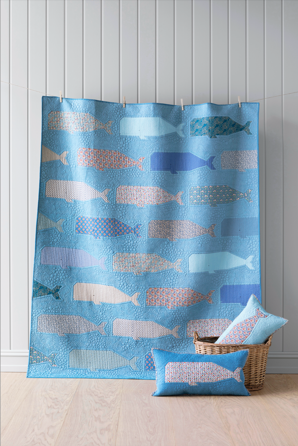 A blue fabric quilt with a repeating pattern of stylized fish in various shades of blue, pink, and beige, hanging in front of a white paneled wall. A matching blue throw pillow and a woven basket with pillows are placed on the light wood floor in front of the curtain.
Photo by Tildafabrics.com [additional_image_link]