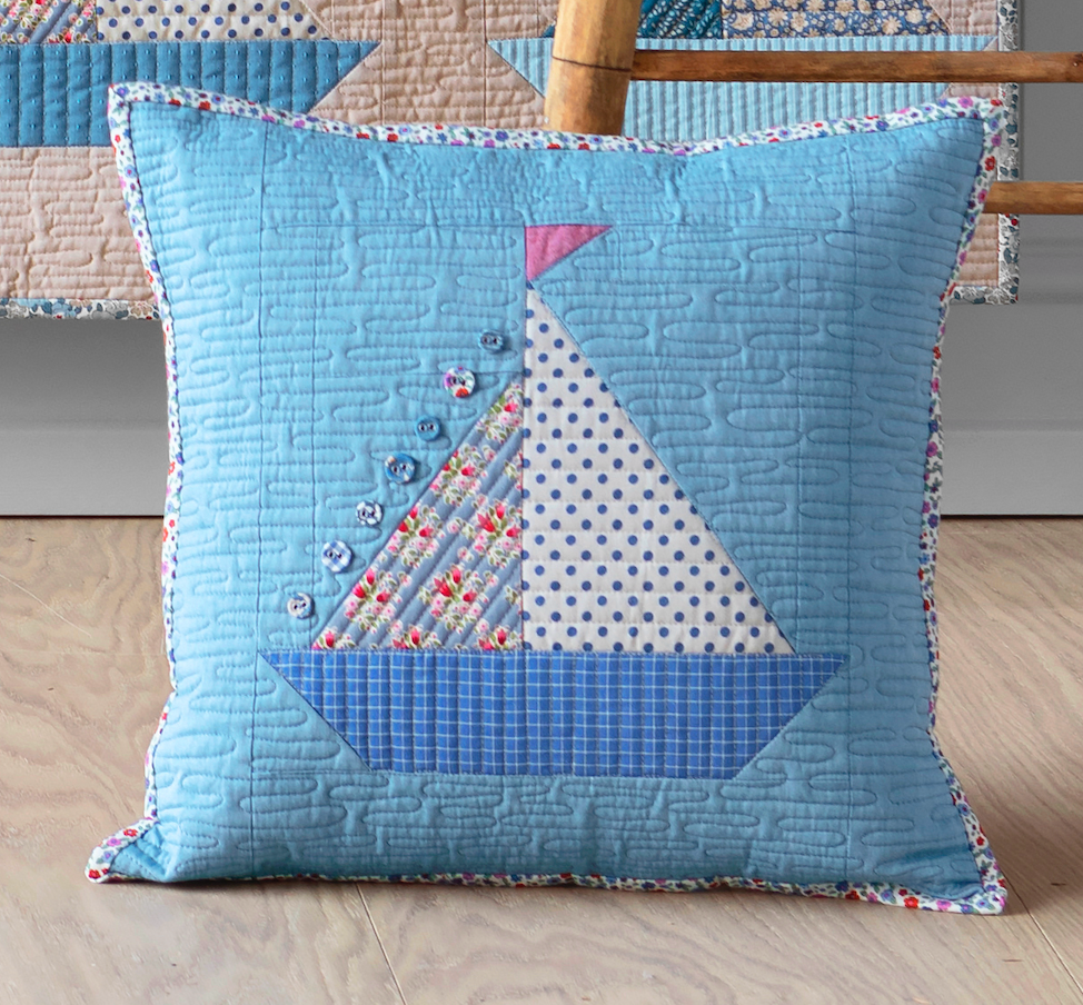 A square quilted throw pillow in light blue with a sailboat patchwork design, displayed on a light wood floor in front of a matching quilt.
Photo by Tildafabrics.com [image_link]