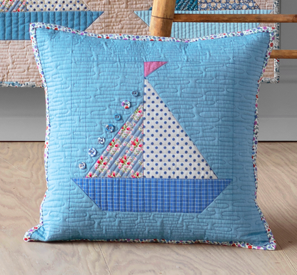 A square quilted throw pillow in light blue with a sailboat patchwork design, displayed on a light wood floor in front of a matching quilt.
Photo by Tildafabrics.com [image_link]