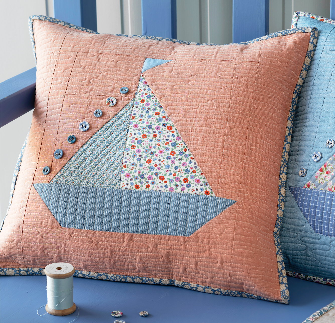 A quilted decorative throw pillow featuring a sailboat patchwork design in peach, light blue, and a multicolor floral pattern, styled on a blue bench next to another pillow.
Photo by Tildafabrics.com [additional_image_link]