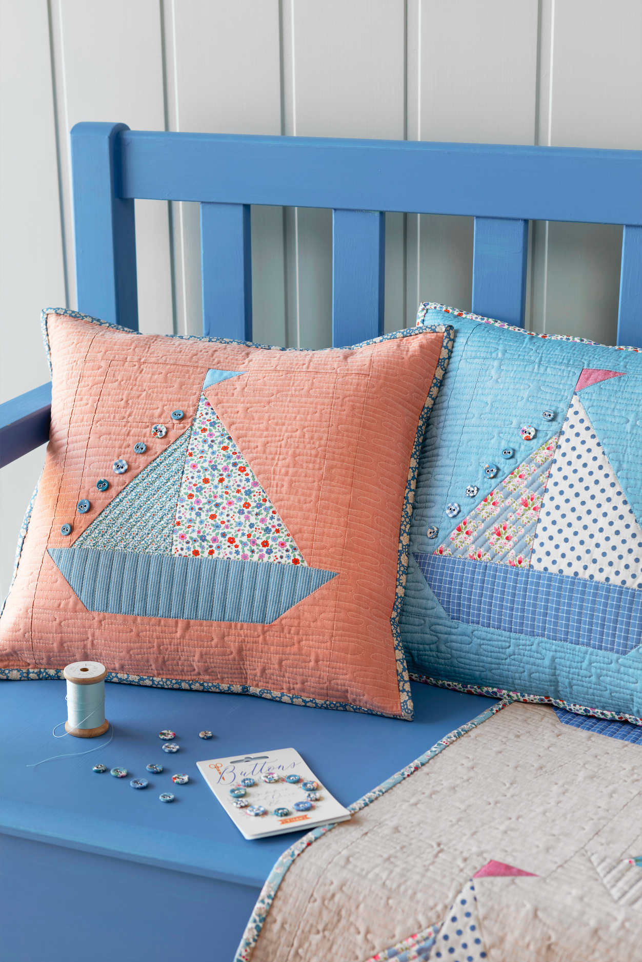 Two decorative quilted throw pillows with sailboat motifs displayed on a blue wooden bench. The pillow on the left is peach with light blue and floral patchwork sailboats, and the pillow on the right is light blue with white and blue patchwork sailboats.
Photo by Tildafabrics.com [additional_image_link]