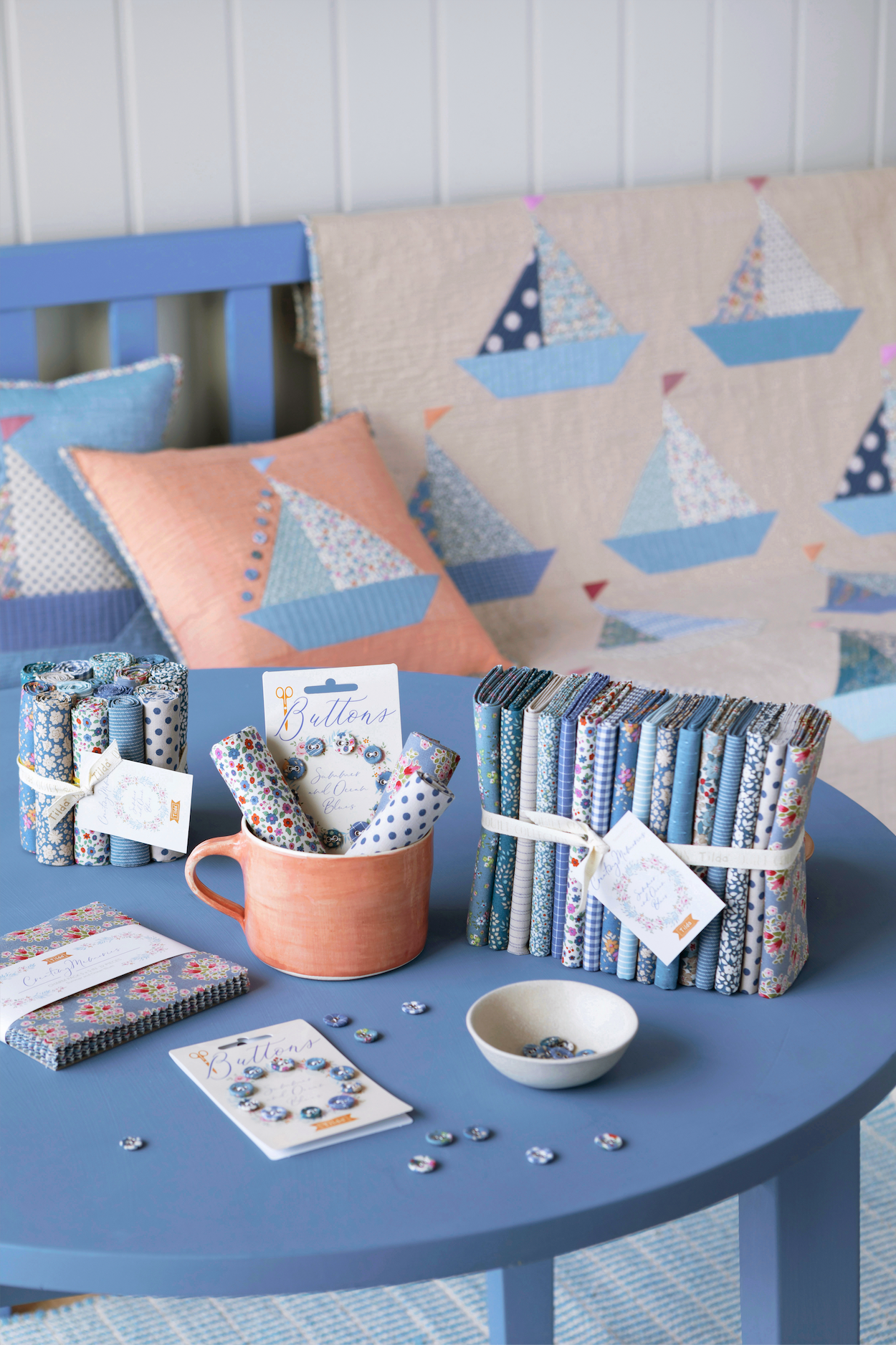 A styled tabletop display of fabric bundles, notebooks, a mug with patterned paper, and small accessories in coordinated blue, navy, white, and coral tones, set on a blue table with a sailboat-patterned quilt and pillows in the background.
Photo by Tildafabrics.com [additional_image_link]