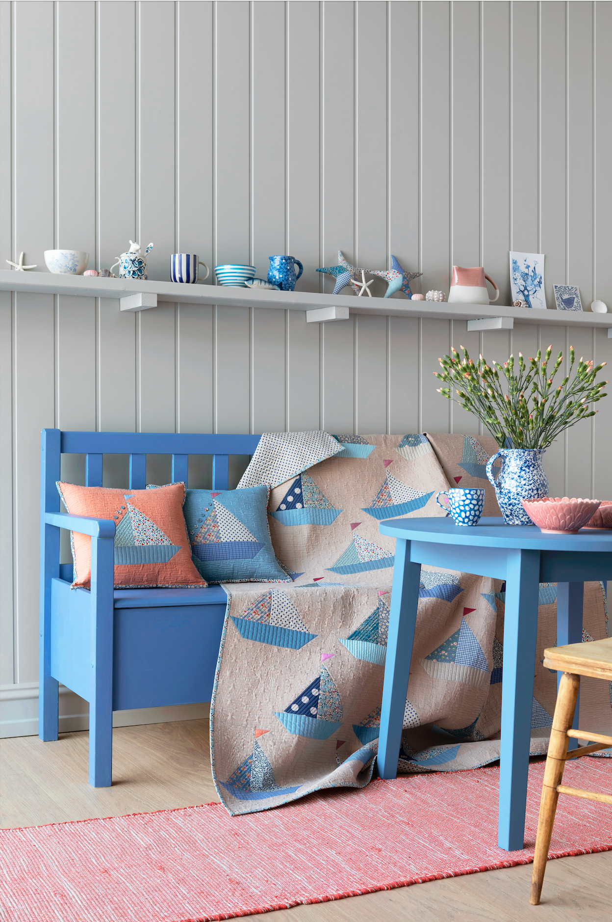 A styled interior scene with a blue wooden bench and matching table, draped with a nautical-patterned quilt. The room has gray paneled walls, a white floating shelf with decorative items, a pink rug, and coral pillows on the bench.
Photo by Tildafabrics.com [additional_image_link]