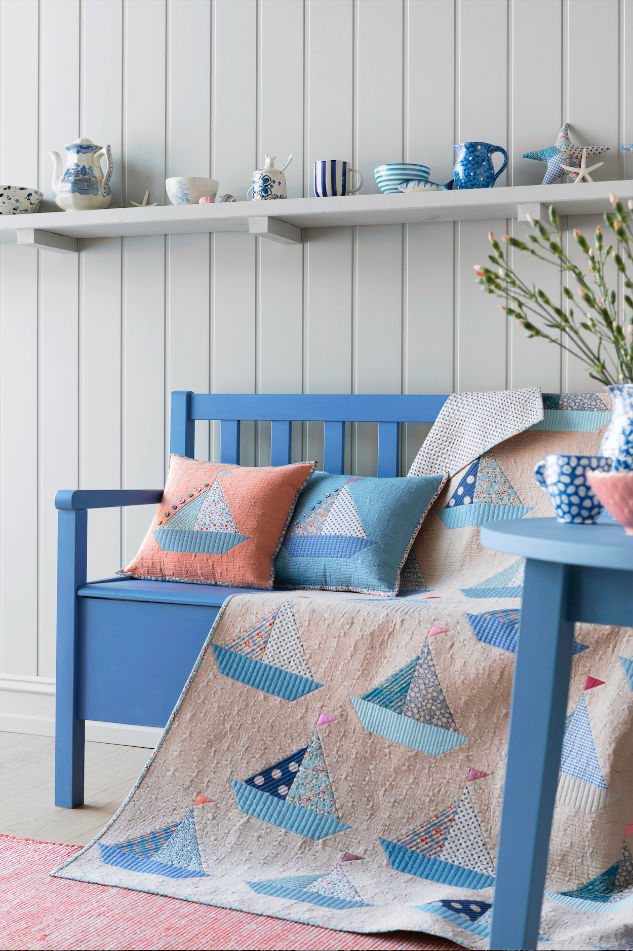 A styled interior scene with a blue wooden bench covered in a beige quilt featuring blue sailboat patterns, accented with matching blue and peach throw pillows. The background features a white paneled wall with a floating shelf holding decorative blue and white ceramic items.
Photo by Tildafabrics.com [additional_image_link]