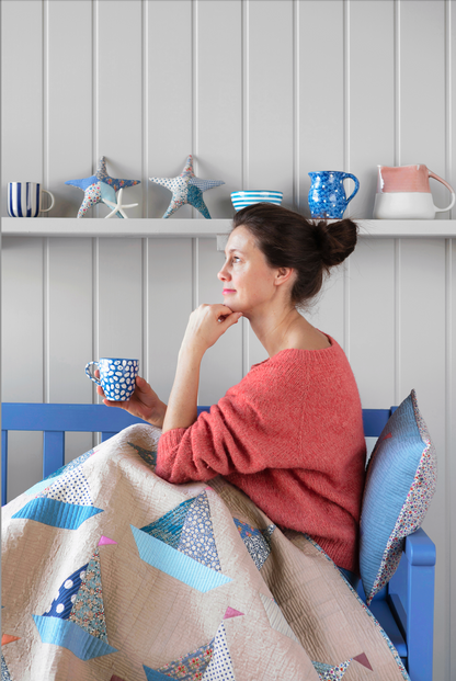 A woman sits in a blue wooden chair wrapped in a patchwork quilt with blue sailboat motifs, holding a blue patterned mug. She is in a coastal-style room with a white paneled wall, a shelf with starfish and mugs, and a blue pillow behind her.
Photo by Tildafabrics.com [additional_image_link]