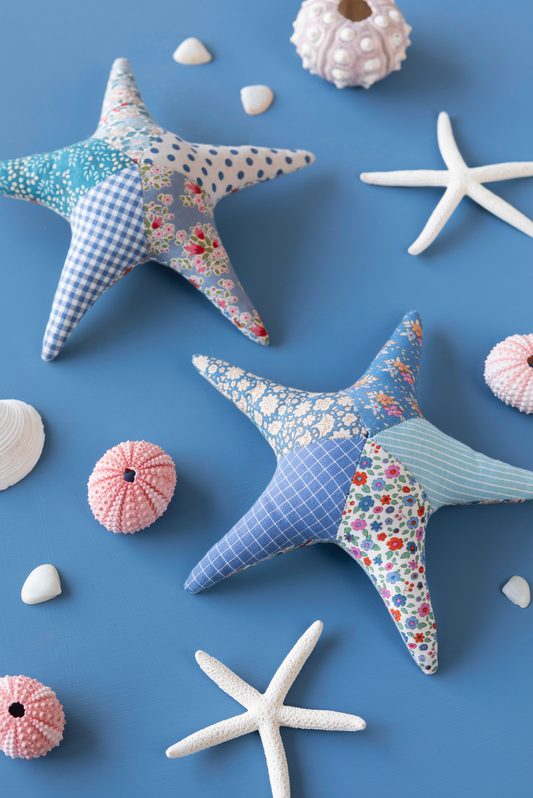 Two handmade patchwork fabric starfish decorations displayed on a blue background with white and pink seashells. The starfish feature various fabric patterns including gingham, floral prints, and stripes in shades of blue, pink, and white.
Photo by Tildafabrics.com [image_link]