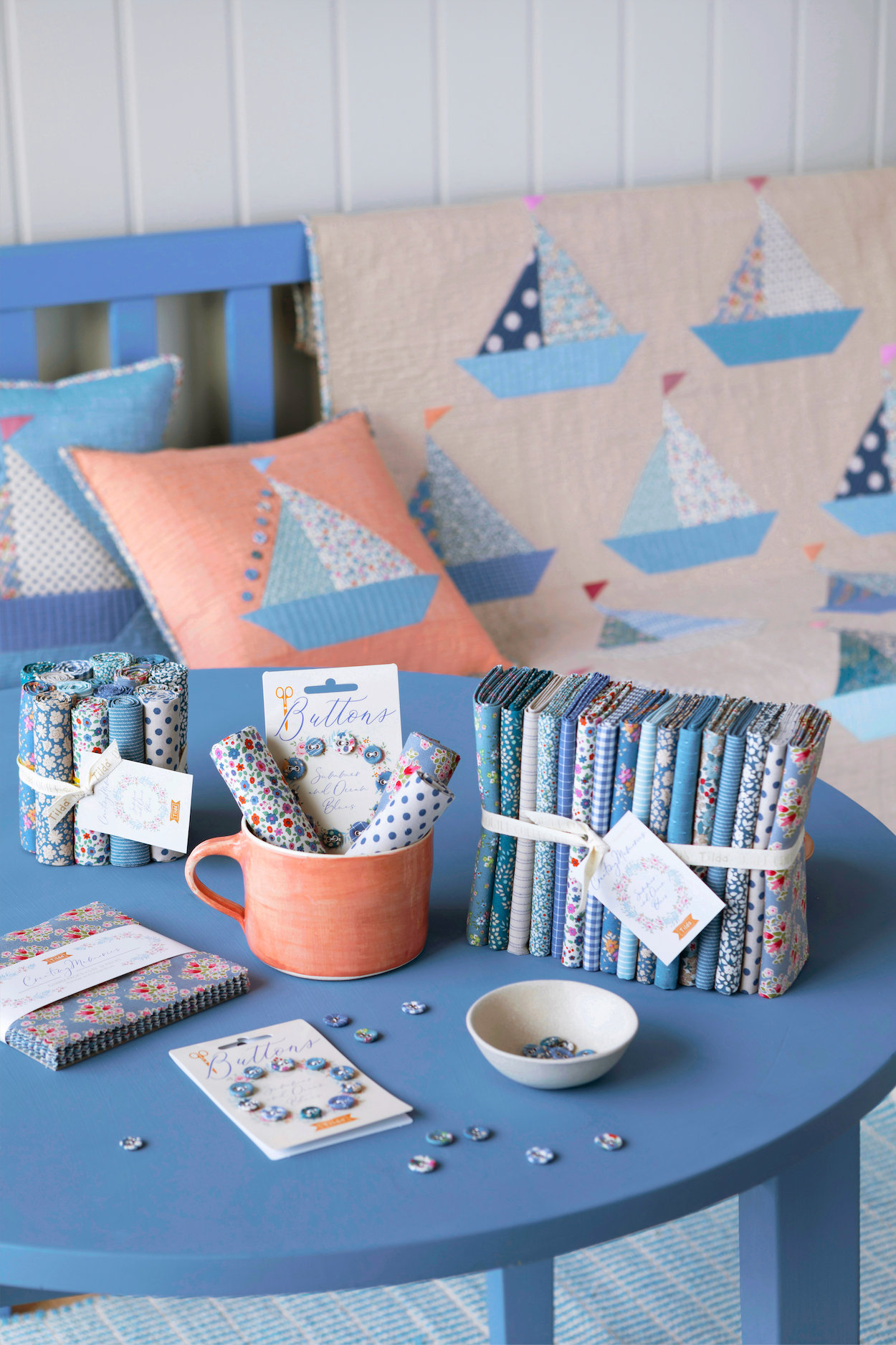 A styled tabletop display of fabric bundles, notebooks, a mug with patterned paper, and small accessories in coordinated blue, navy, white, and coral tones, set on a blue table with a sailboat-patterned quilt and pillows in the background.
Photo by Tildafabrics.com [additional_image_link]