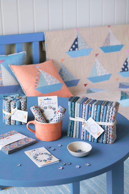 A styled tabletop display of fabric bundles, notebooks, a mug with patterned paper, and small accessories in coordinated blue, navy, white, and coral tones, set on a blue table with a sailboat-patterned quilt and pillows in the background.
Photo by Tildafabrics.com [additional_image_link]