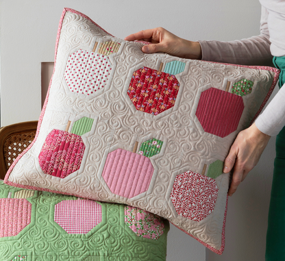 A quilted throw pillow with apple motifs in pink, red, and green fabrics, held by a person wearing a beige top and green pants. The pillow features a cream-colored quilted swirl background and is layered over another matching quilted pillow on a chair.
Photo by Tildafabrics.com [image_link]