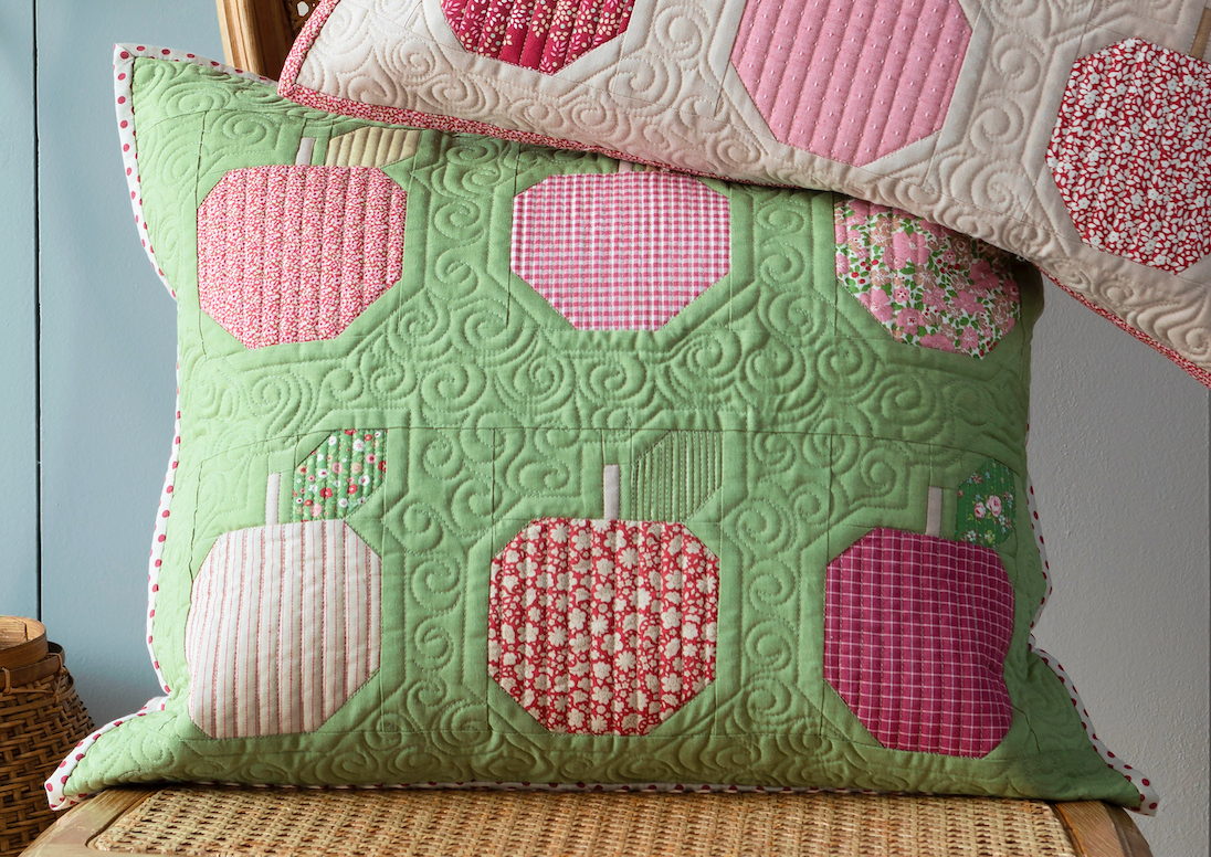 A quilted rectangular throw pillow in light green with patchwork apple motifs in pink, red, and cream, styled on a woven chair with a second matching pillow partially visible behind it.
Photo by Tildafabrics.com [additional_image_link]
