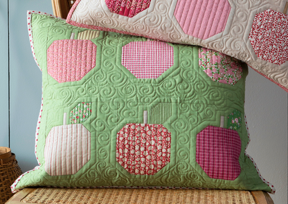 A quilted rectangular throw pillow in light green with patchwork apple motifs in pink, red, and cream, styled on a woven chair with a second matching pillow partially visible behind it.
Photo by Tildafabrics.com [additional_image_link]